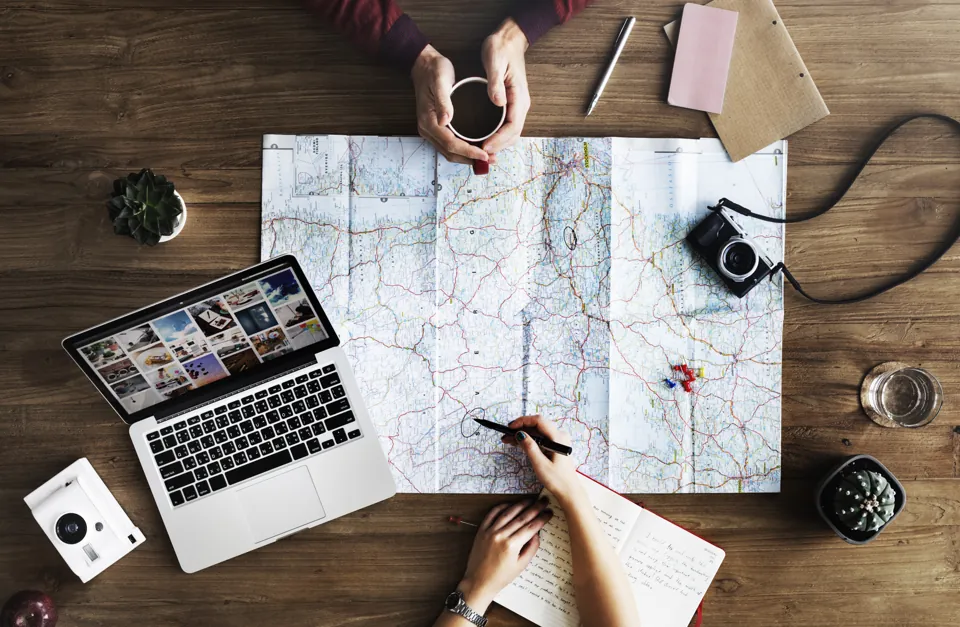 Manos de dos personas preparando un viaje con mapa, libreta y computadora