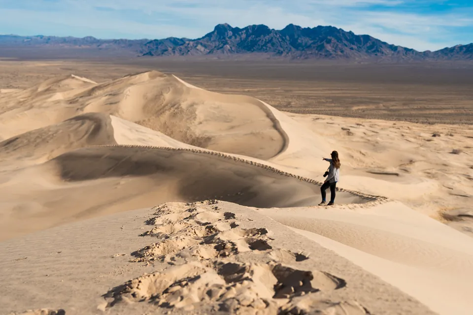 Persona sobre una duna de arena con el desierto y montañas al fondo en Kelso Dunes.